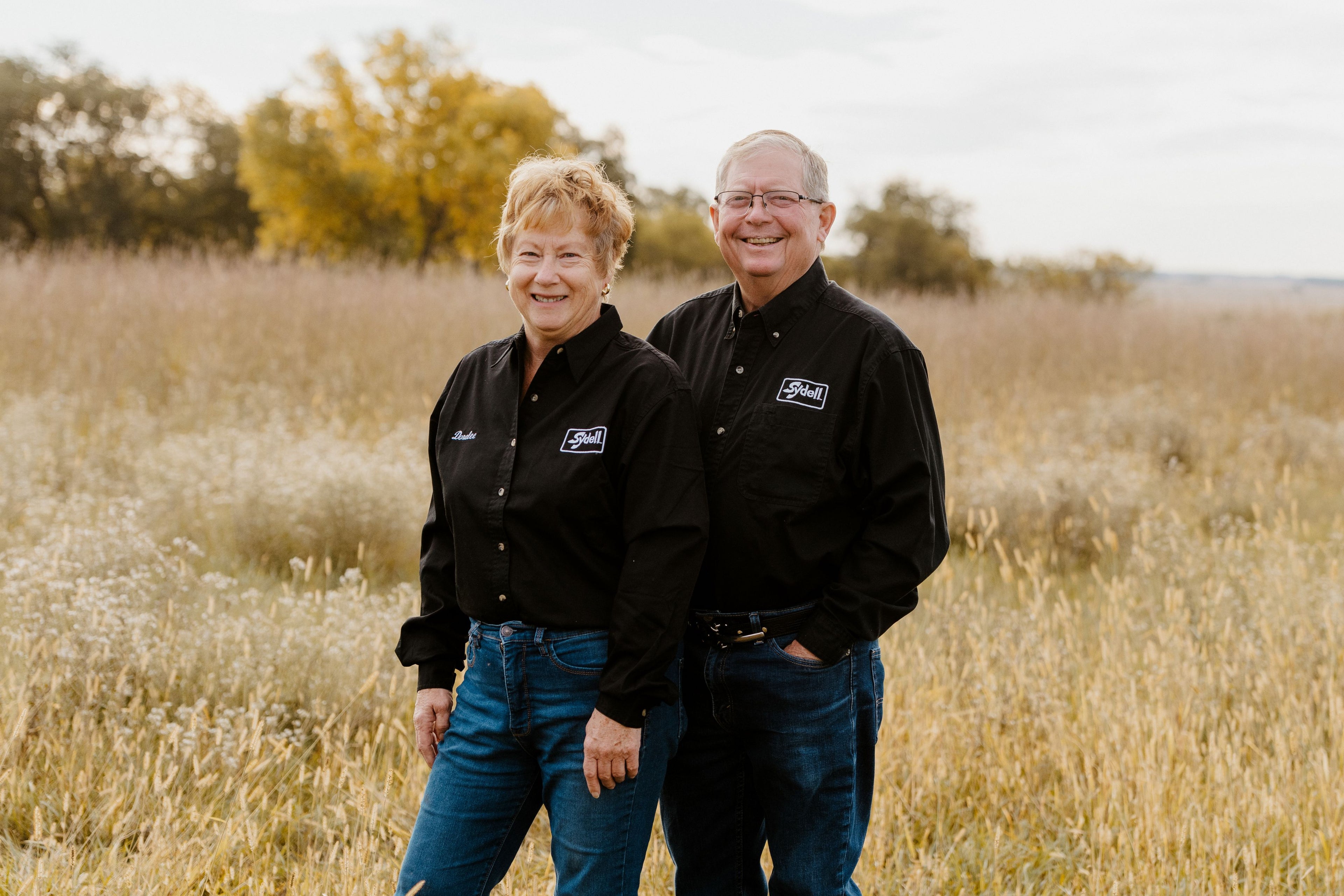 Two people standing in a field wearing matching black shirts with Sydell logos